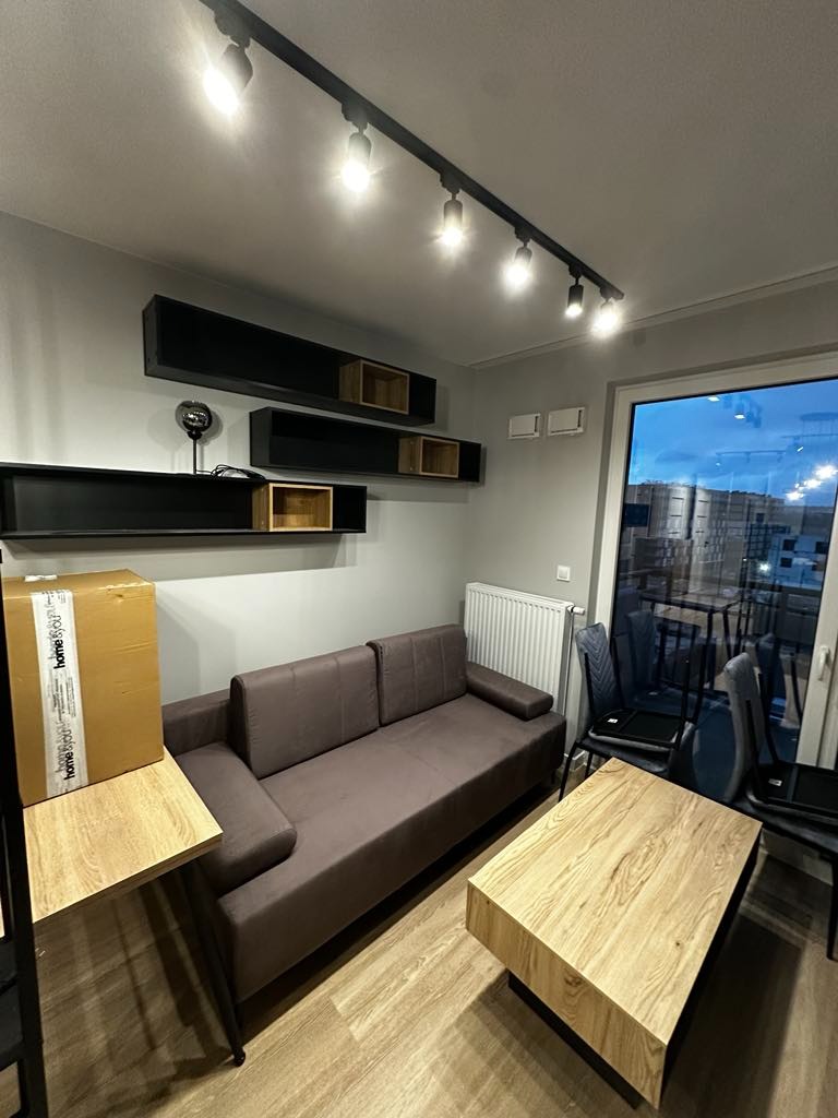 Nowoczesny salon z szarą ścianą, drewnianą podłogą, brązową sofą, drewnianym stolikiem kawowym, czarnymi półkami wiszącymi z drewnianymi wstawkami, oświetleniem szynowym i kartonem na stole, widok...