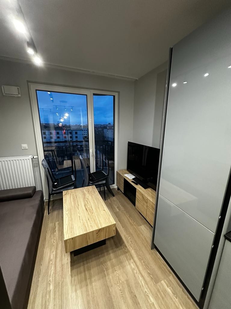 Jasne panele podłogowe w nowoczesnym salonie z drewnianym stolikiem kawowym, szarą sofą i szafką RTV pod telewizorem, widok na miasto z balkonu.