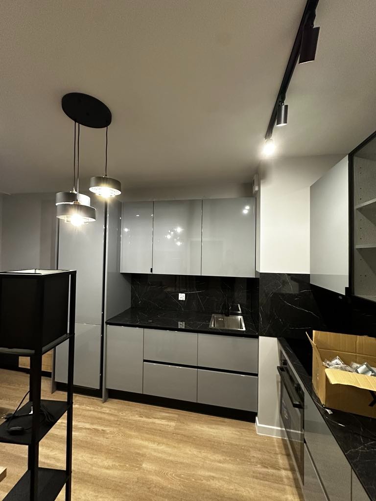 Nowoczesna kuchnia z szarymi, lakierowanymi frontami szafek, czarnym blatem imitującym marmur, oświetleniem szynowym i wiszącymi lampami nad wyspą, częściowo rozpakowana z kartonami na blacie.