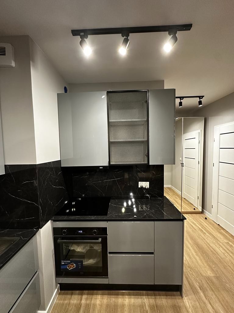 Nowoczesna kuchnia z szarymi szafkami, czarnym blatem imitującym marmur i drewnopodobną podłogą; oświetlenie punktowe sufitowe.