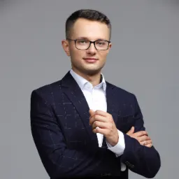 Portret mężczyzny w okularach i granatowej marynarce w kratę, z rękami skrzyżowanymi na piersiach, na jednolitym szarym tle.