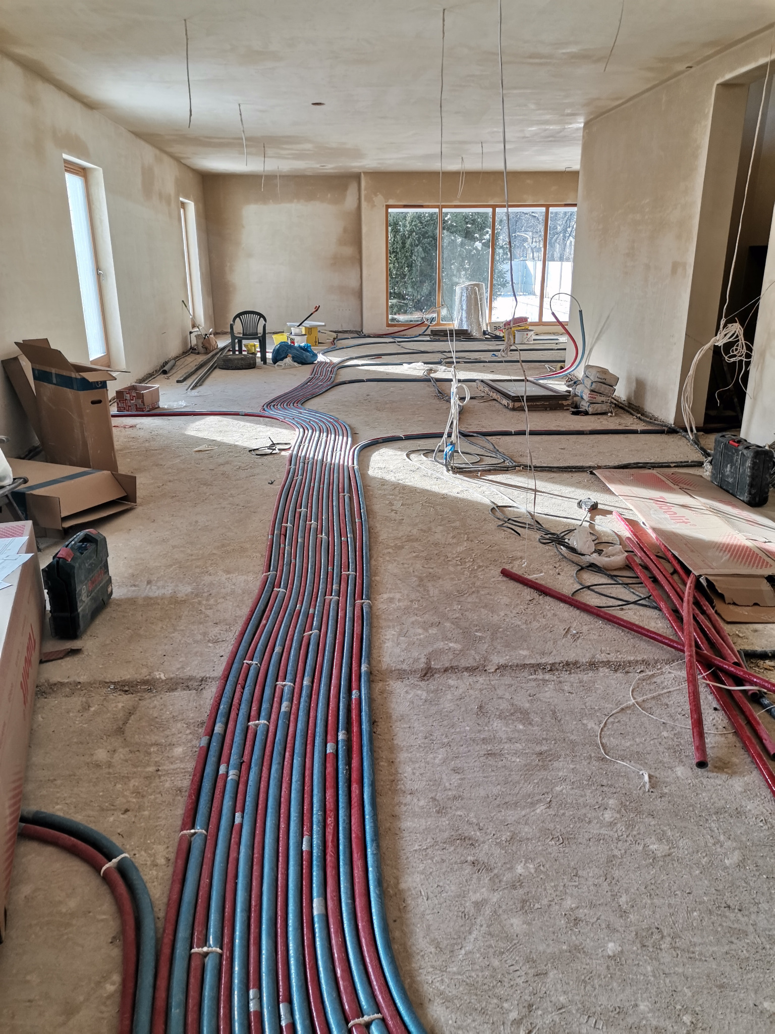 Rozłożone rury ogrzewania podłogowego w surowym wnętrzu, widoczne okna z zimowym krajobrazem, przygotowania do wylewki.