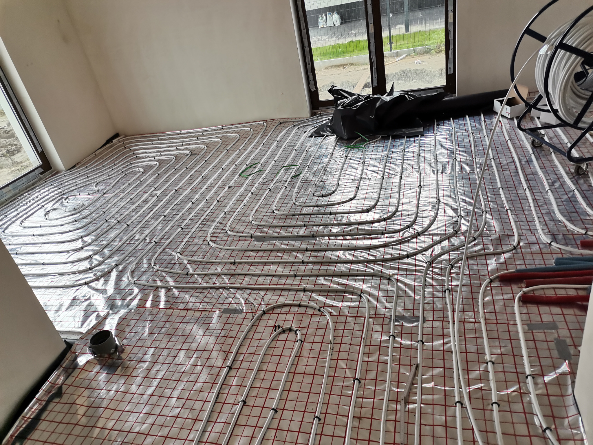 Rozłożone rury ogrzewania podłogowego na folii z siatką, widoczne okna i zwój rur na wózku w tle.