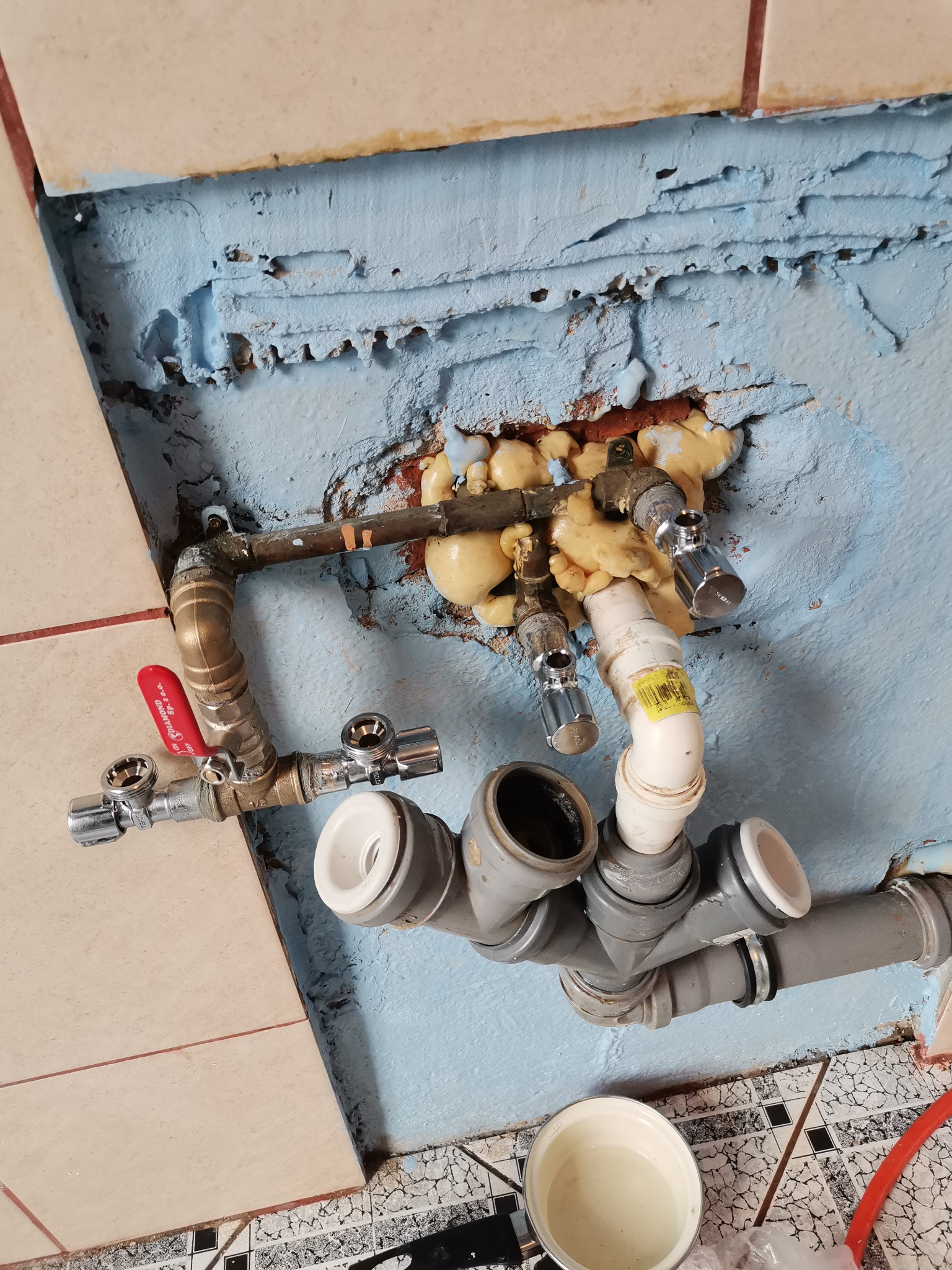 Połączenie rur miedzianych i plastikowych z zaworami, zabezpieczone pianką montażową na tle niebieskiej ściany z częściowo położonymi płytkami.