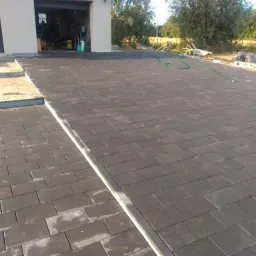 Ułożona, prostokątna kostka brukowa w kolorze grafitowym przed garażem, z widocznymi krawężnikami i przygotowanym podłożem pod schody.