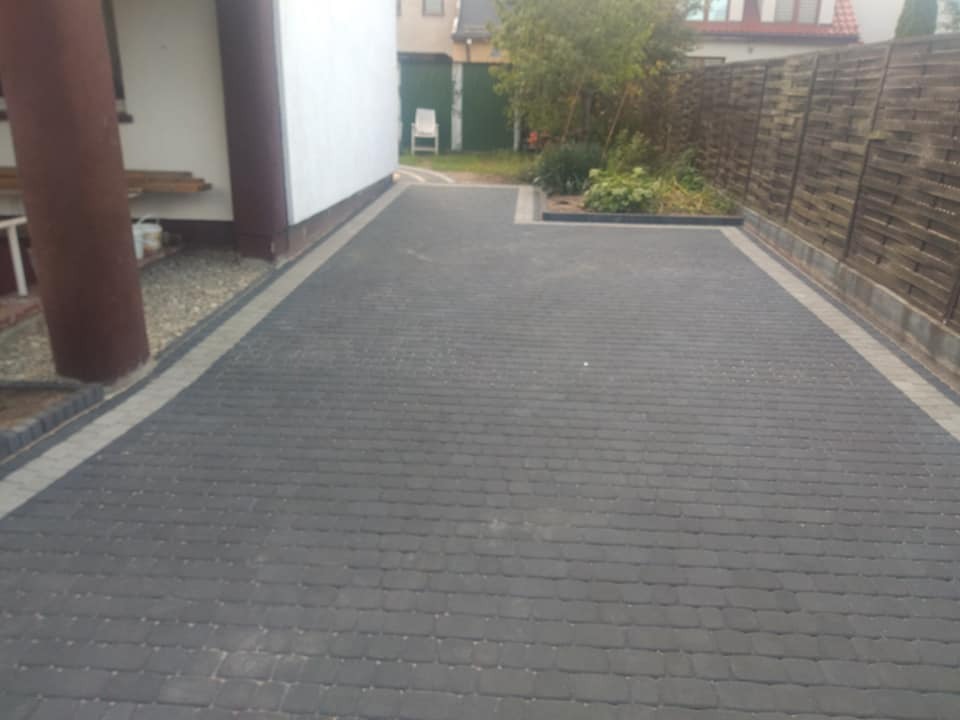 Ułożona, ciemnoszara kostka brukowa prowadząca od budynku do drewnianego ogrodzenia. Obrzeża wykończone jaśniejszą kostką, widoczne rośliny i elementy architektury ogrodowej.