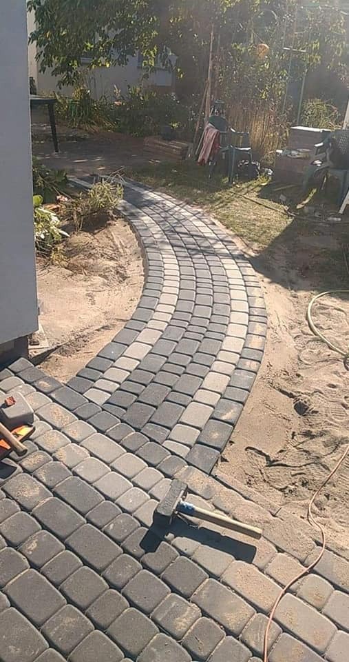 Ścieżka z dwukolorowej kostki ułożona w łuk, z widocznym gumowym młotkiem i piaskiem, prowadząca przez trawnik w słoneczny dzień.