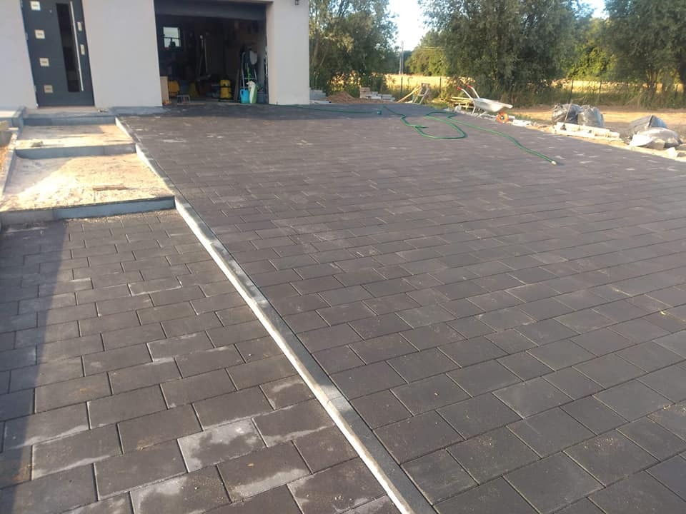 Ułożona, prostokątna kostka brukowa w kolorze grafitowym przed garażem, z widocznymi krawężnikami i przygotowanym podłożem pod schody.