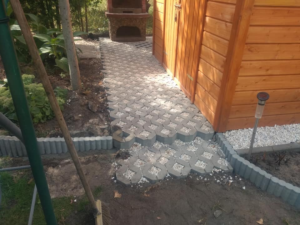 Ułożona szara ażurowa ścieżka z kostki brukowej prowadząca do drewnianej altany, obrzeża z szarego betonu, w tle grill ogrodowy z kamienia.