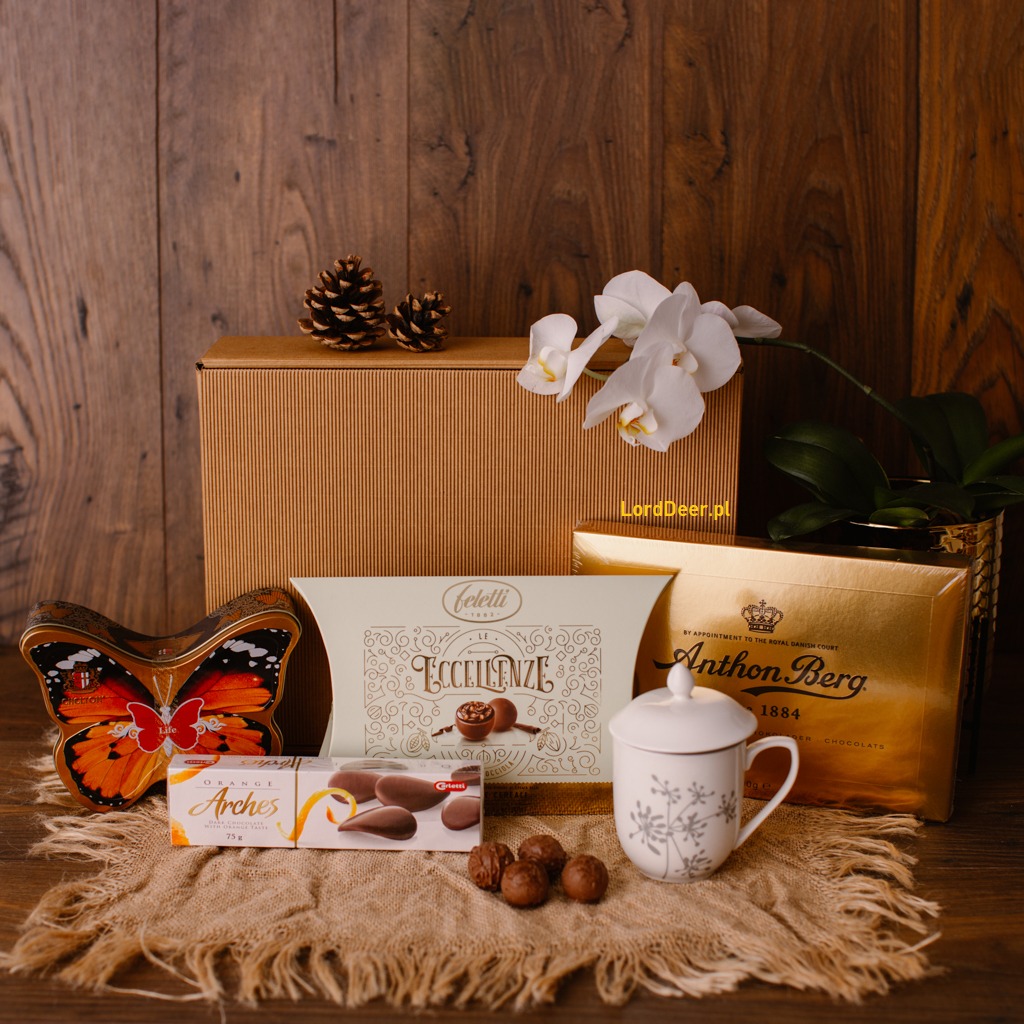 Zestaw upominkowy: pudełko z szyszkami, czekoladki Feletti Eccellenze, Anthon Berg, Arches, puszka w kształcie motyla i filiżanka z przykrywką na jutowej serwetce, na tle drewnianej ściany.