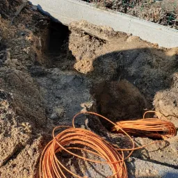 Wykop w ziemi przy ogrodzeniu z widocznym pomarańczowym przewodem zwiniętym w krąg, prawdopodobnie przygotowanie do instalacji odgromowej.