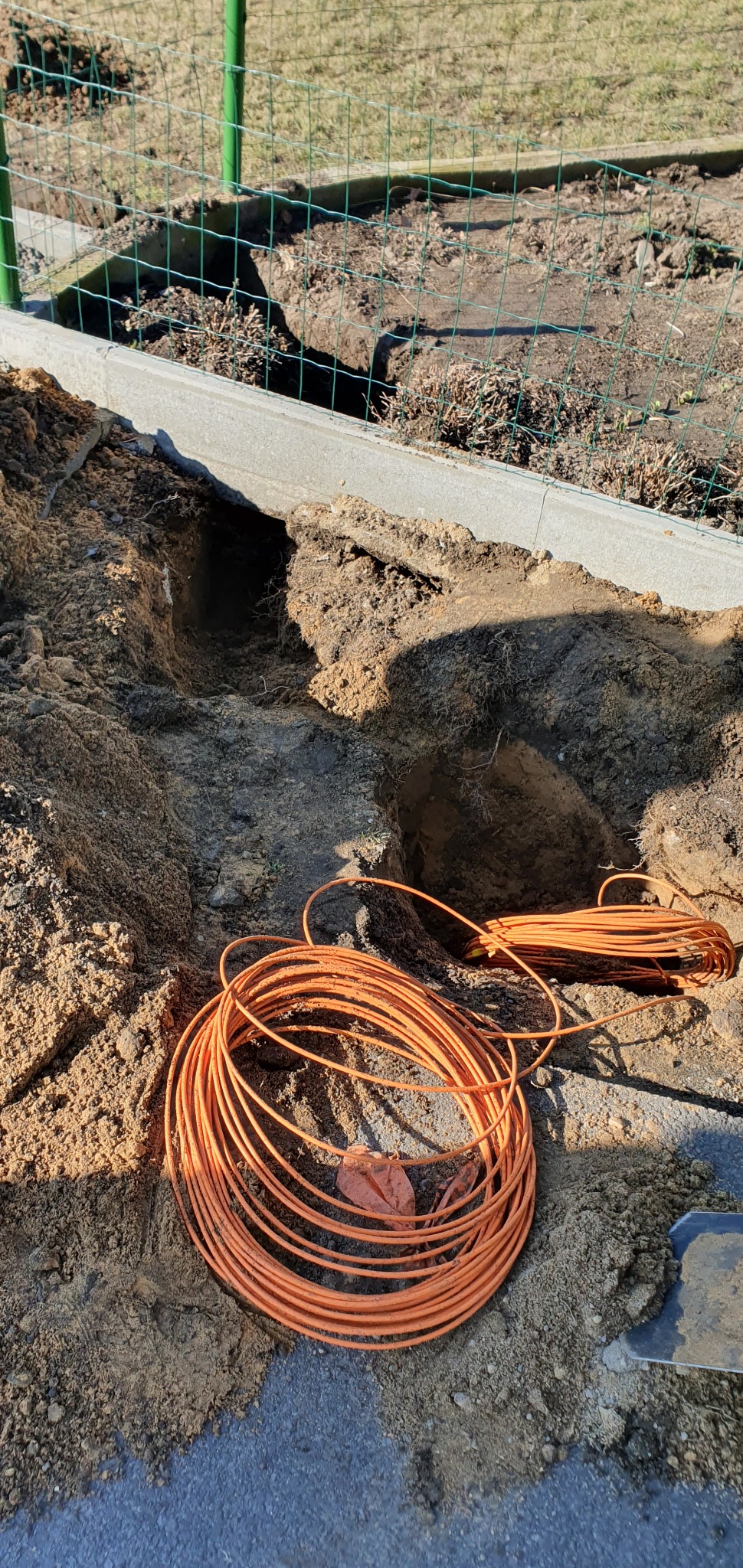 Wykop w ziemi przy ogrodzeniu z widocznym pomarańczowym przewodem zwiniętym w krąg, prawdopodobnie przygotowanie do instalacji odgromowej.