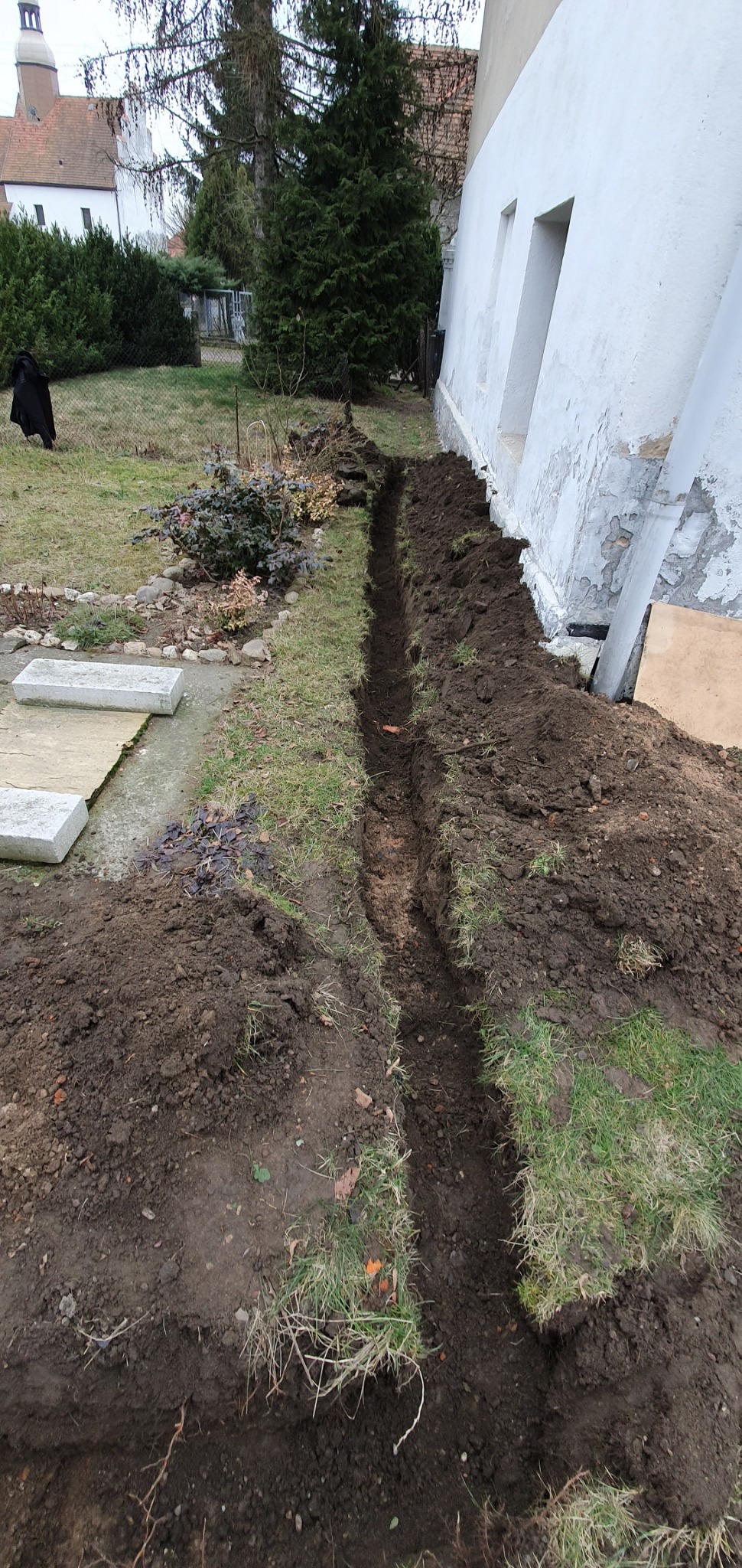 Wykopany wąski rów wzdłuż ściany budynku z widocznymi fragmentami instalacji i zniszczonym tynkiem, przygotowany prawdopodobnie pod instalację odgromową.