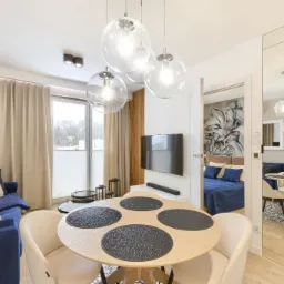 Jasne wnętrze mieszkania z okrągłym stołem z czterema czarnymi podkładkami, niebieską sofą i widokiem na sypialnię przez otwarte drzwi, oświetlone wiszącymi szklanymi lampami.