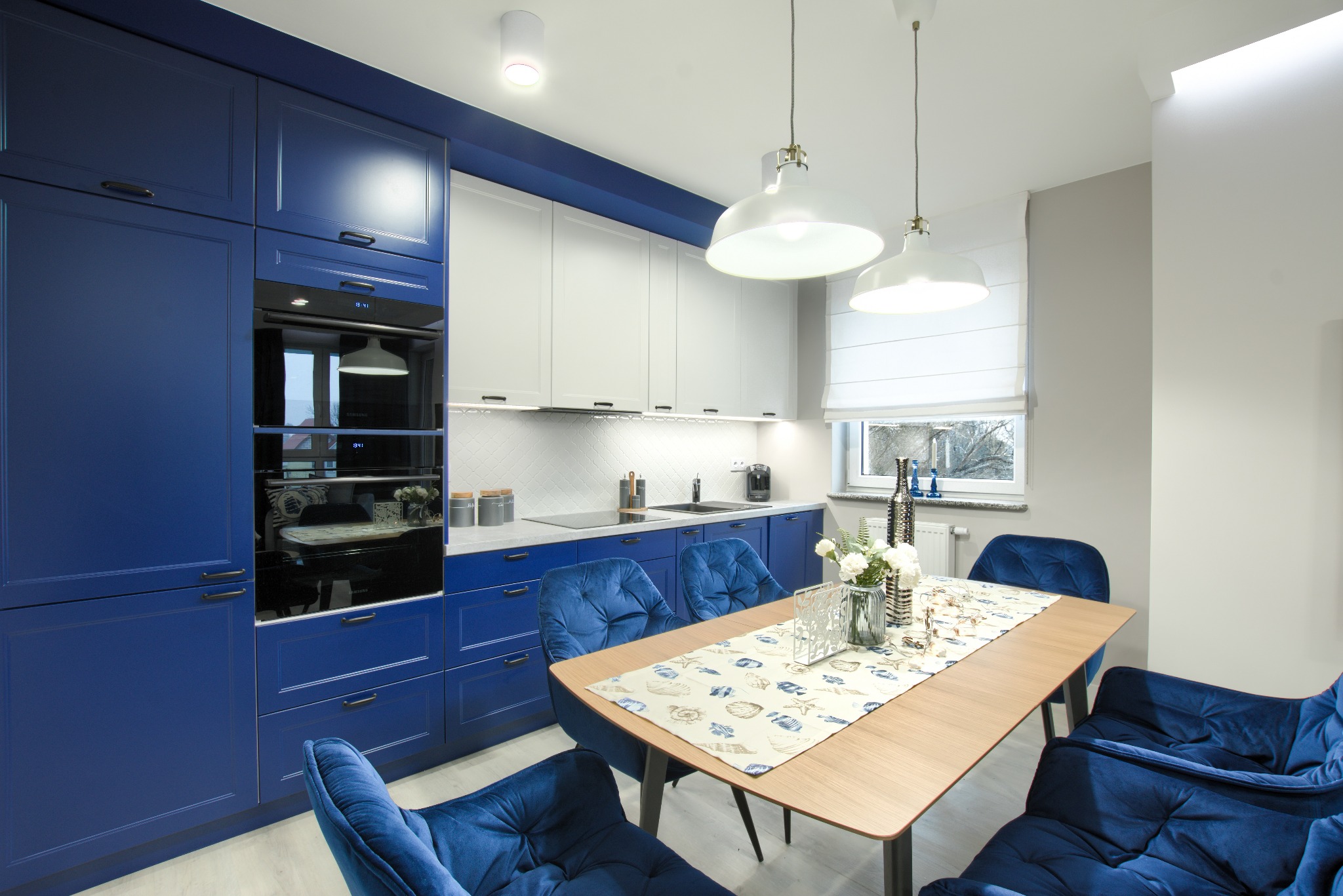 Nowoczesna kuchnia z granatowymi szafkami, białymi górnymi szafkami i drewnianym stołem otoczonym granatowymi krzesłami z aksamitu, oświetlona wiszącymi lampami.