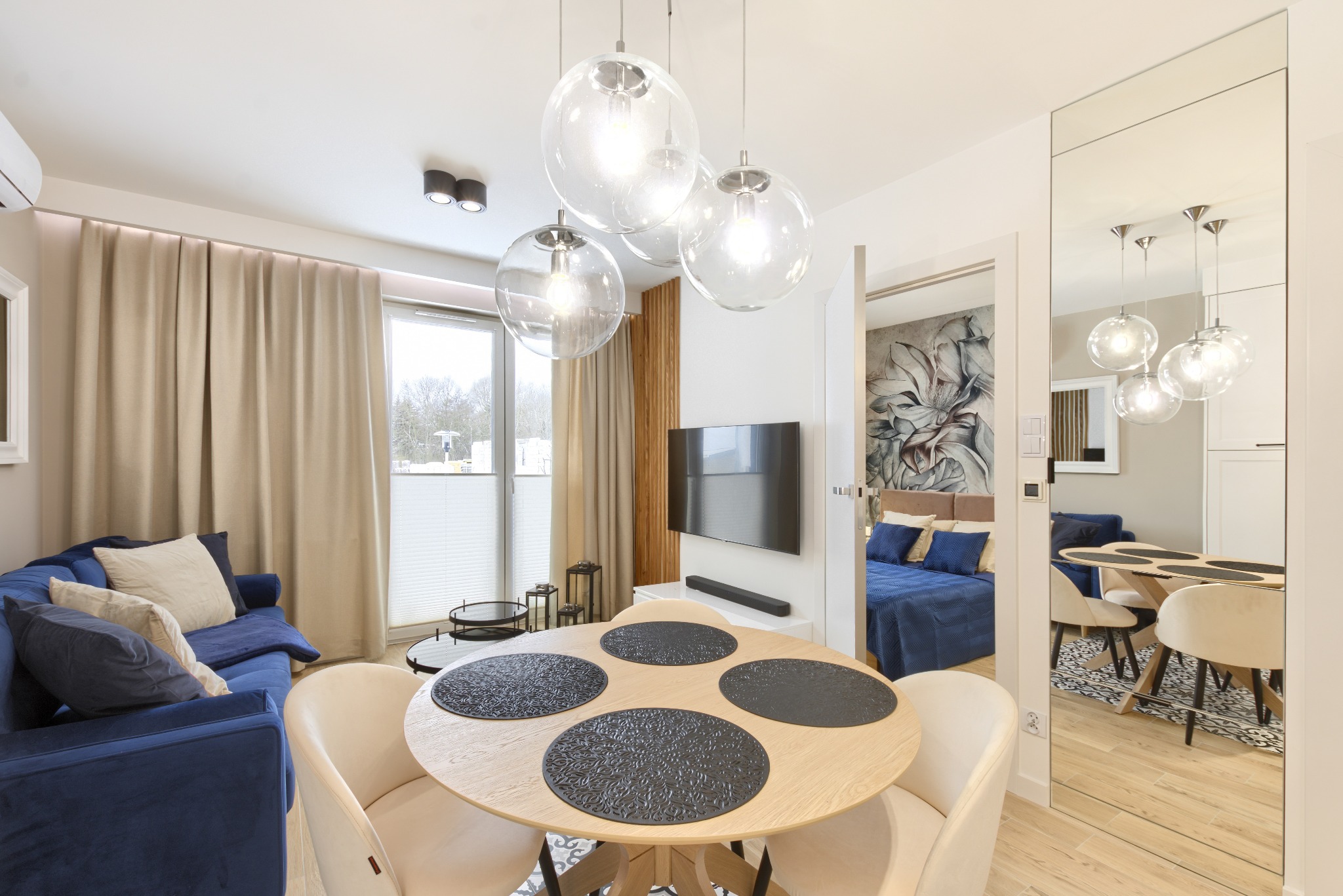 Jasne wnętrze mieszkania z okrągłym stołem z czterema czarnymi podkładkami, niebieską sofą i widokiem na sypialnię przez otwarte drzwi, oświetlone wiszącymi szklanymi lampami.