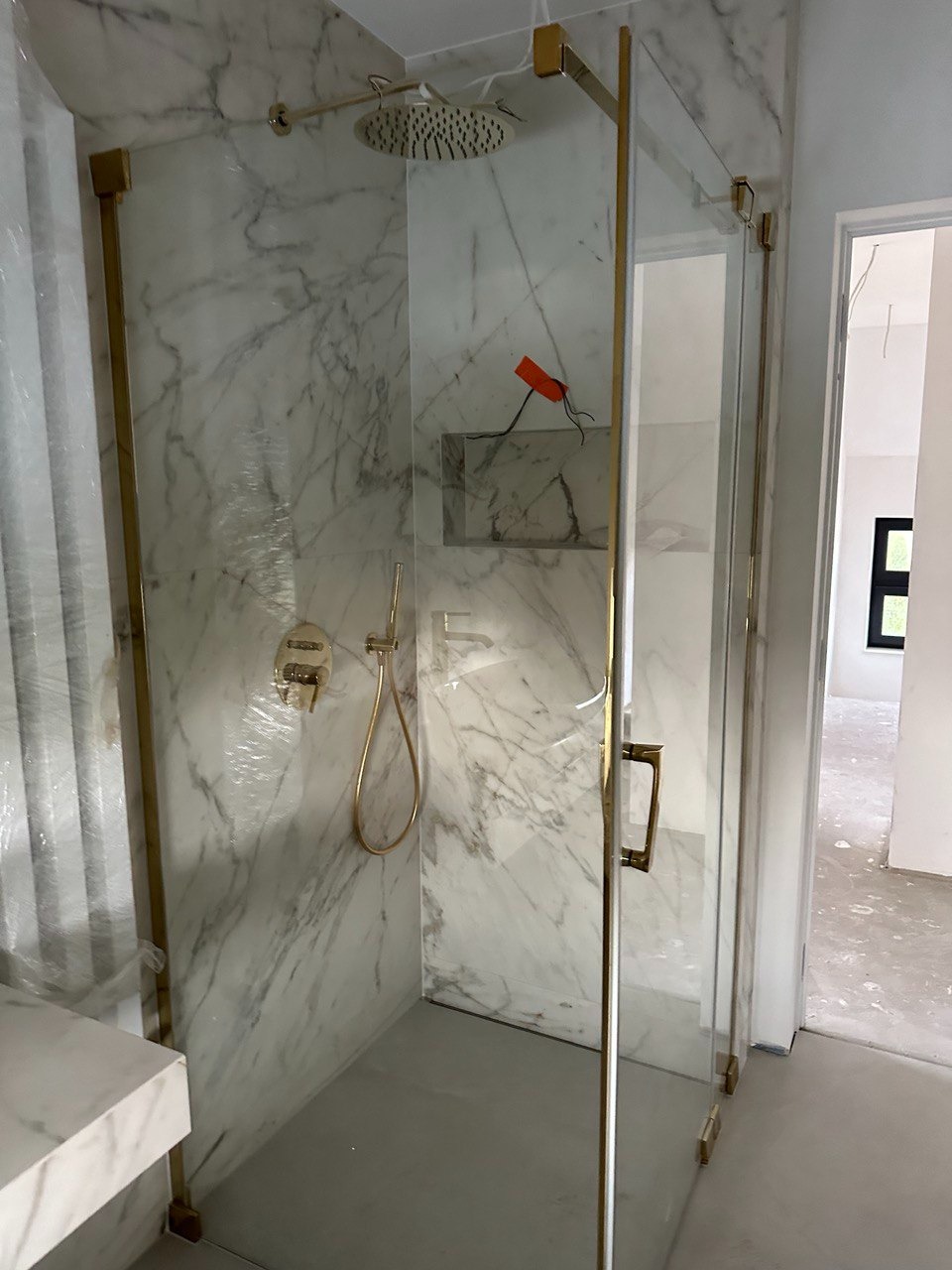 Kabina prysznicowa z marmurowymi ścianami i złotymi okuciami, widoczne elementy armatury i odpływ liniowy, częściowo zabezpieczona folią ochronną.