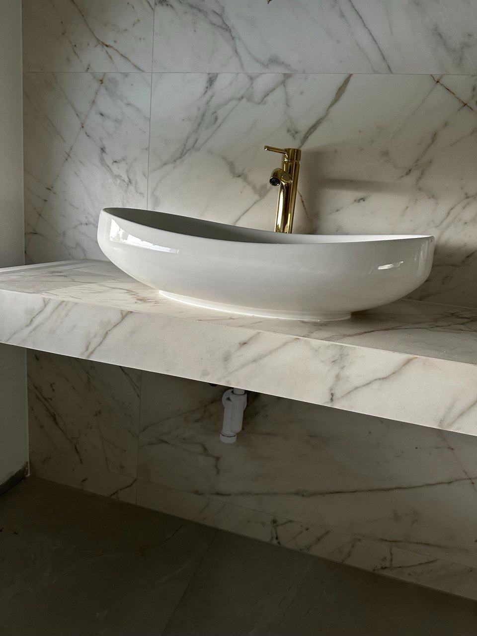 Owalna, biała umywalka nablatowa z złotą baterią na marmurowym blacie i ścianie, widoczny biały syfon.