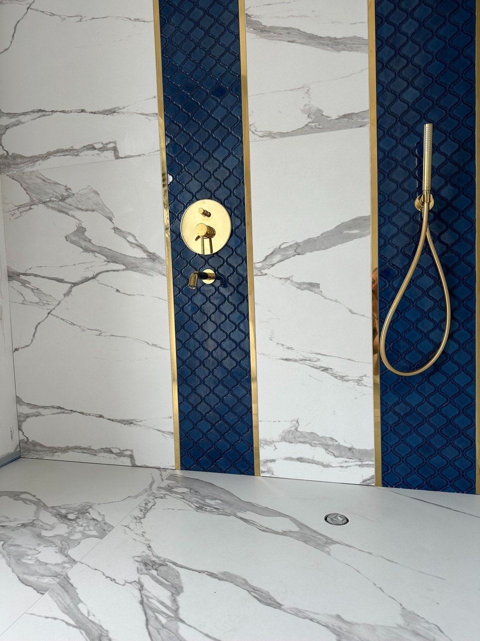 Luksusowa łazienka z marmurowymi płytkami i niebieskimi akcentami, złota armatura prysznicowa i dekoracyjne listwy.