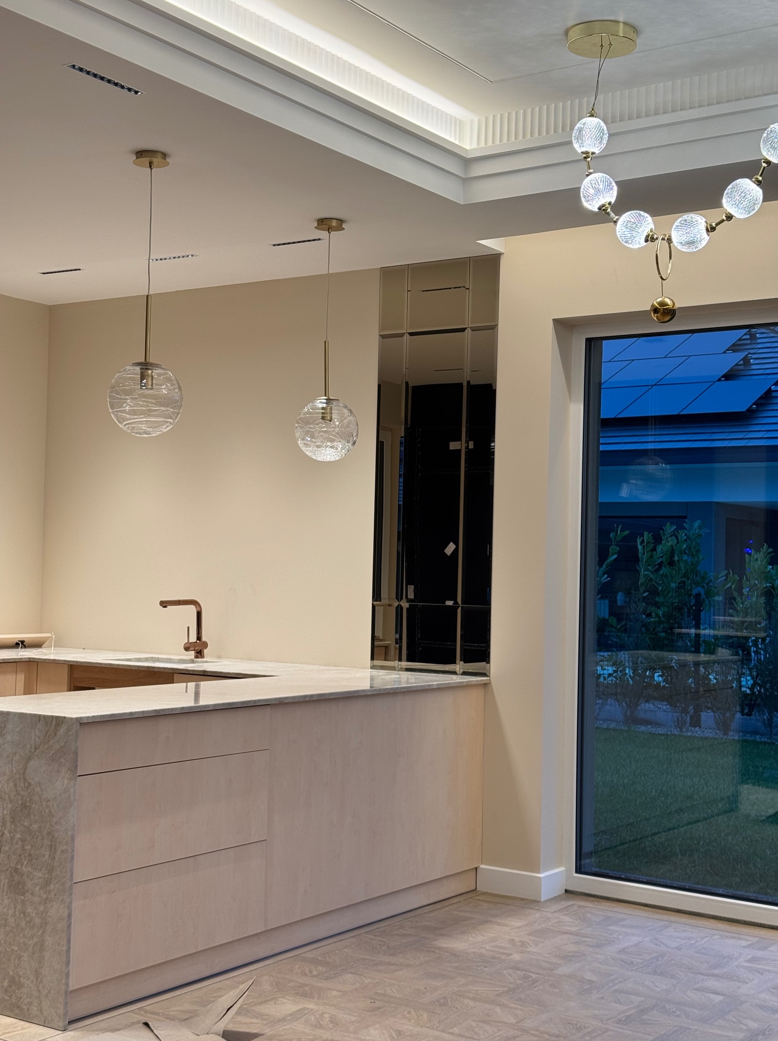 Nowoczesna kuchnia w odcieniach beżu z marmurowym blatem, miedzianą armaturą i dekoracyjnymi lampami wiszącymi, uzupełniona lustrzanymi panelami ściennymi i dużym oknem z widokiem na ogród.