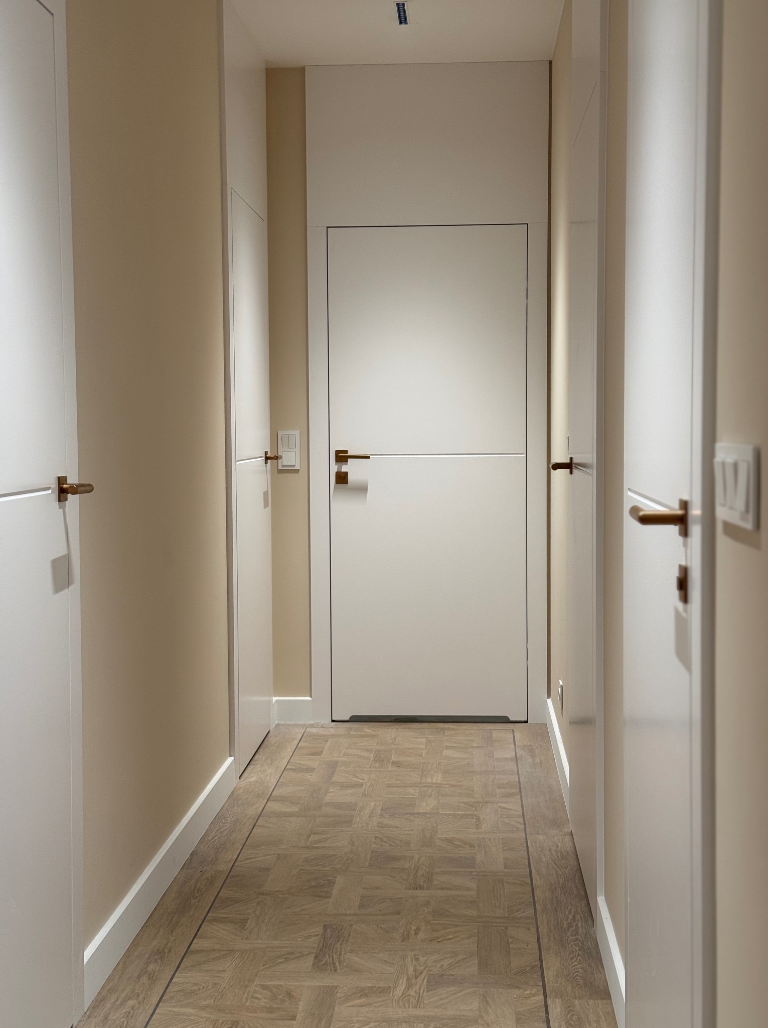 Korytarz z parkietem w jodełkę, białymi drzwiami z poziomym frezem i mosiężnymi klamkami, ściany w kolorze ciepłego beżu.