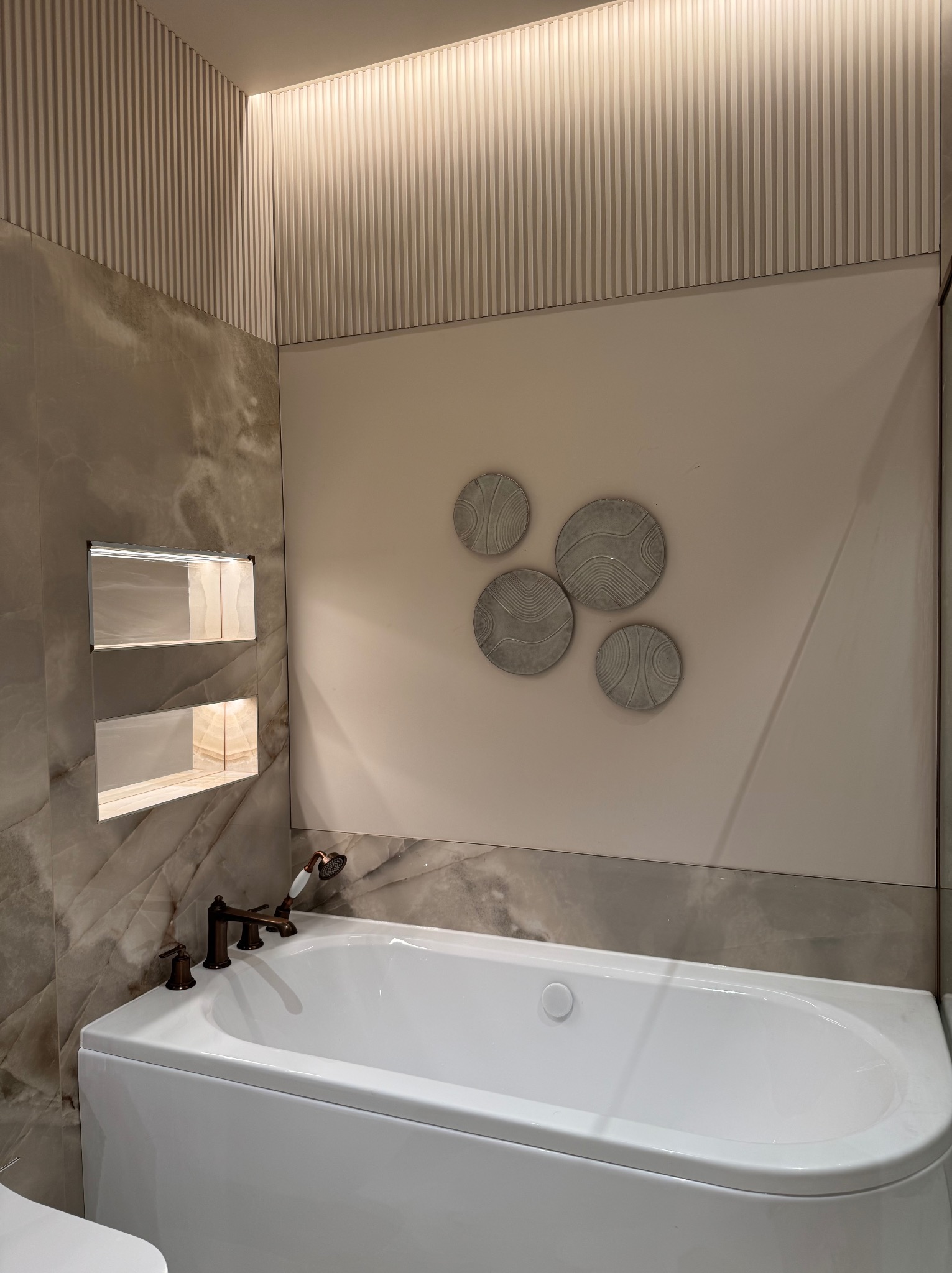 Nowoczesna łazienka z białą wanną, armaturą w kolorze brązu i dekoracyjnymi okrągłymi elementami na ścianie. Ściana wyłożona płytkami imitującymi marmur z wnęką oświetloną od wewnątrz.