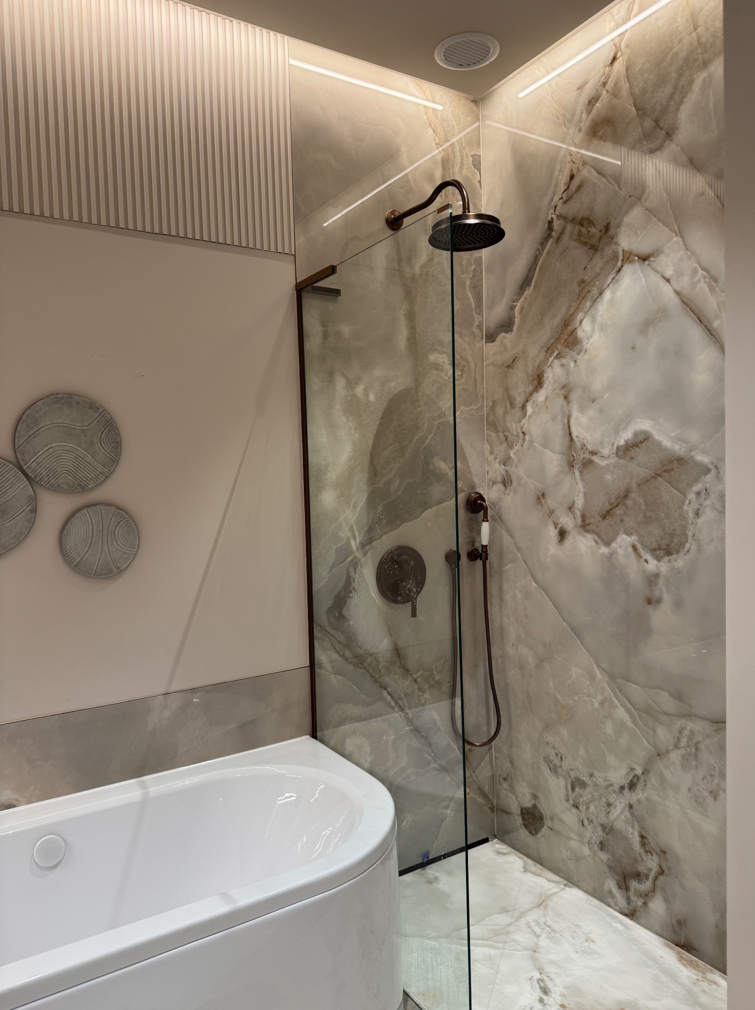 Nowoczesna łazienka z białą wanną, kabiną prysznicową z marmurowymi ścianami, deszczownicą w kolorze miedzi i dekoracyjnymi okrągłymi panelami na ścianie.