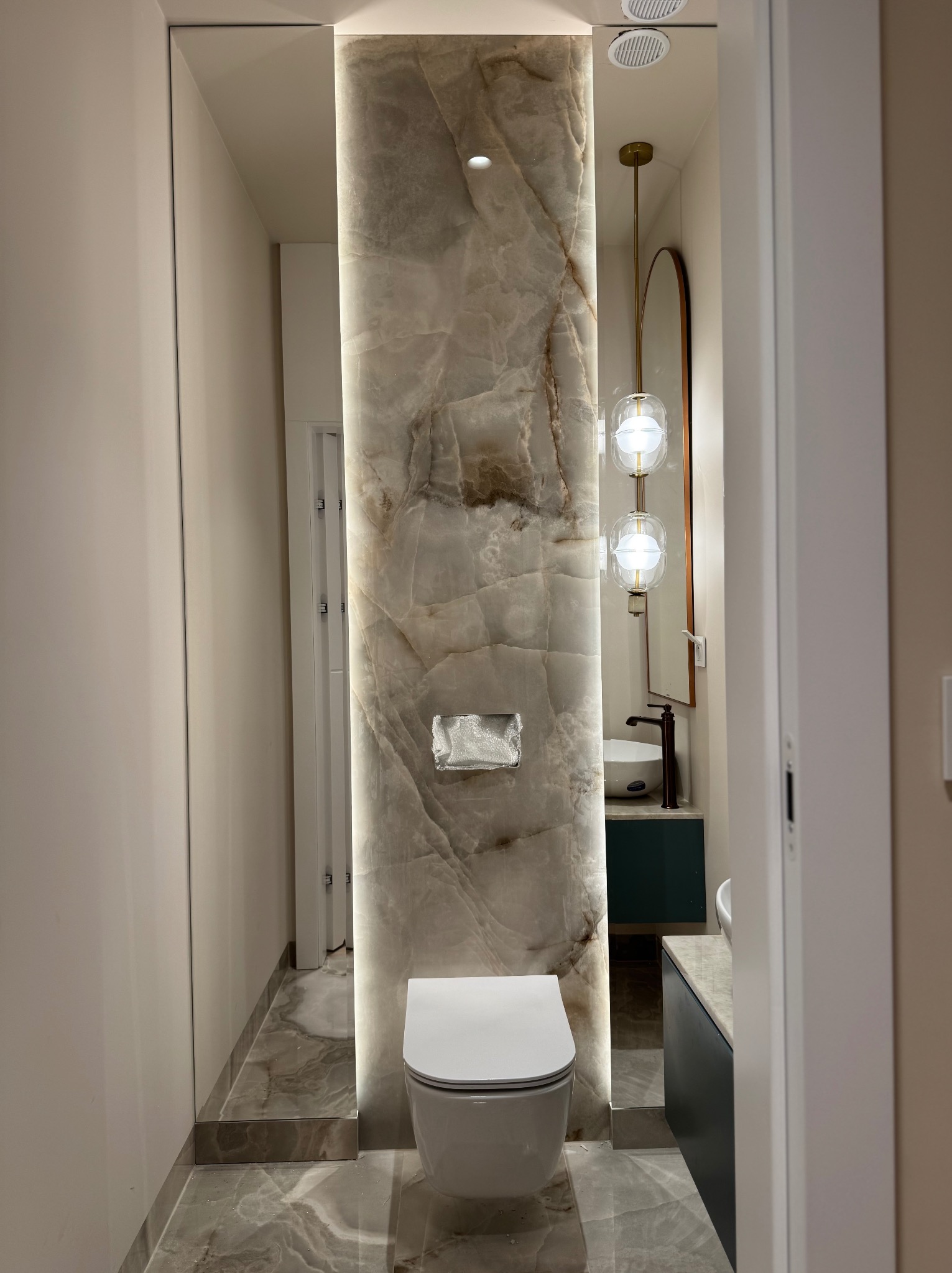 Nowoczesna łazienka z podświetlanym panelem z onyksu, wiszącą toaletą i umywalką z ciemnozieloną szafką oraz okrągłym lustrem.