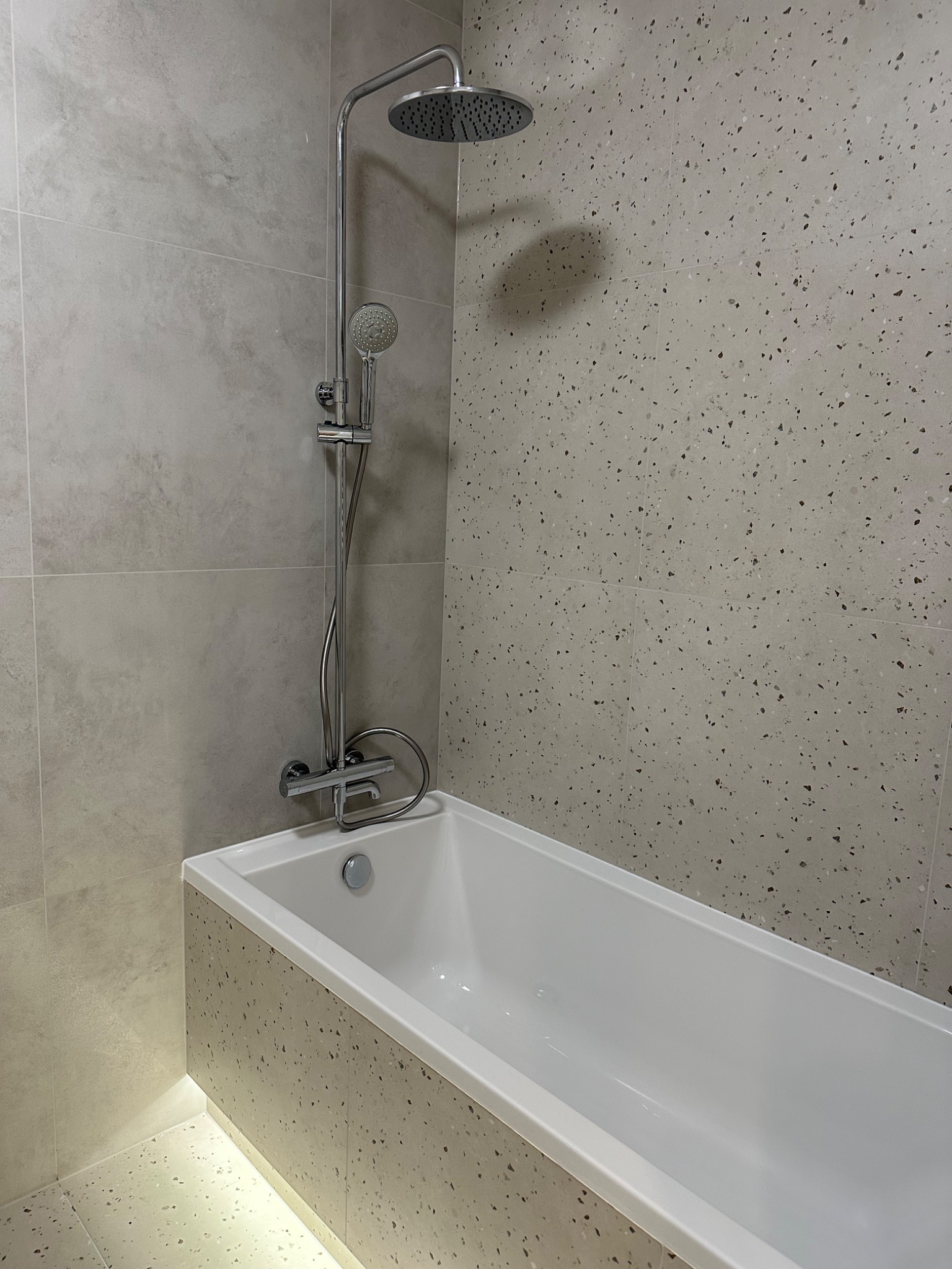 Nowoczesna łazienka z wanną obudowaną płytkami lastryko, chromowana armatura prysznicowa i ściany wyłożone płytkami w odcieniach szarości.