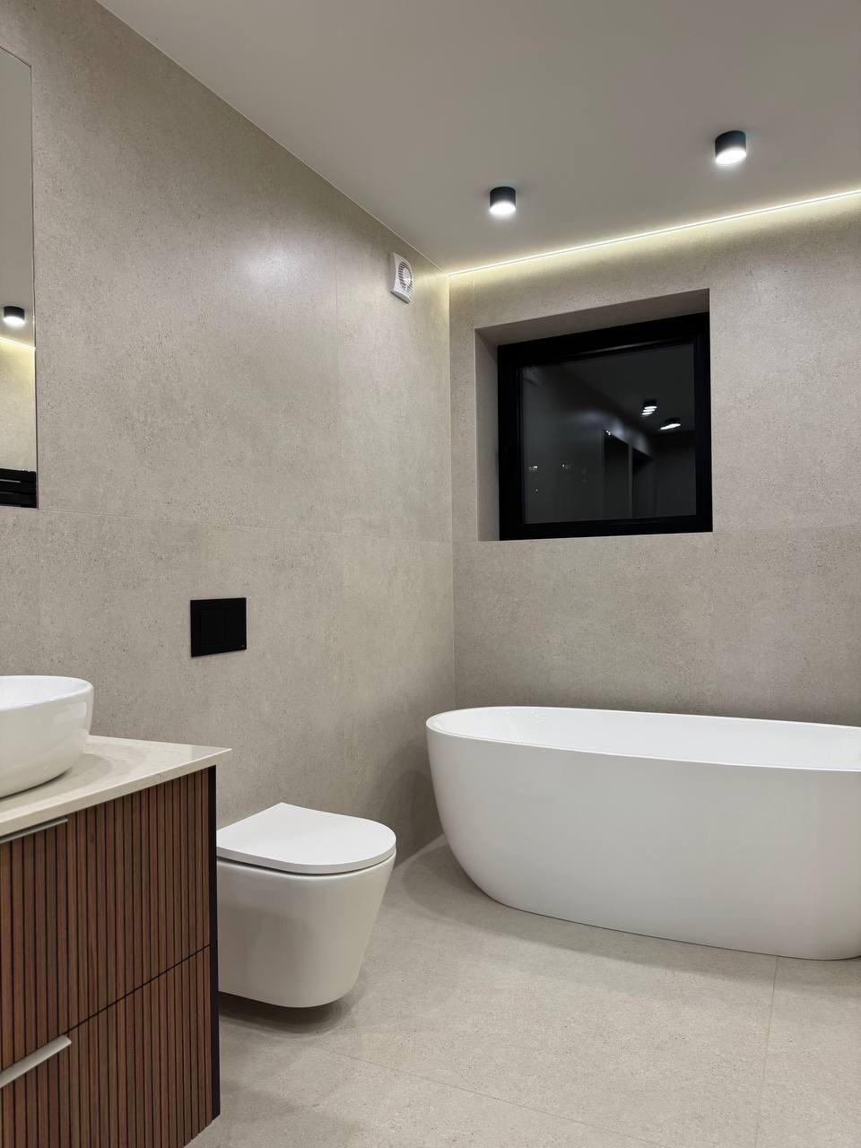 Nowoczesna łazienka z beżowymi płytkami, wolnostojącą białą wanną, podwieszaną toaletą, umywalką nablatową na szafce z drewnianymi lamelami i oknem z czarną ramą.