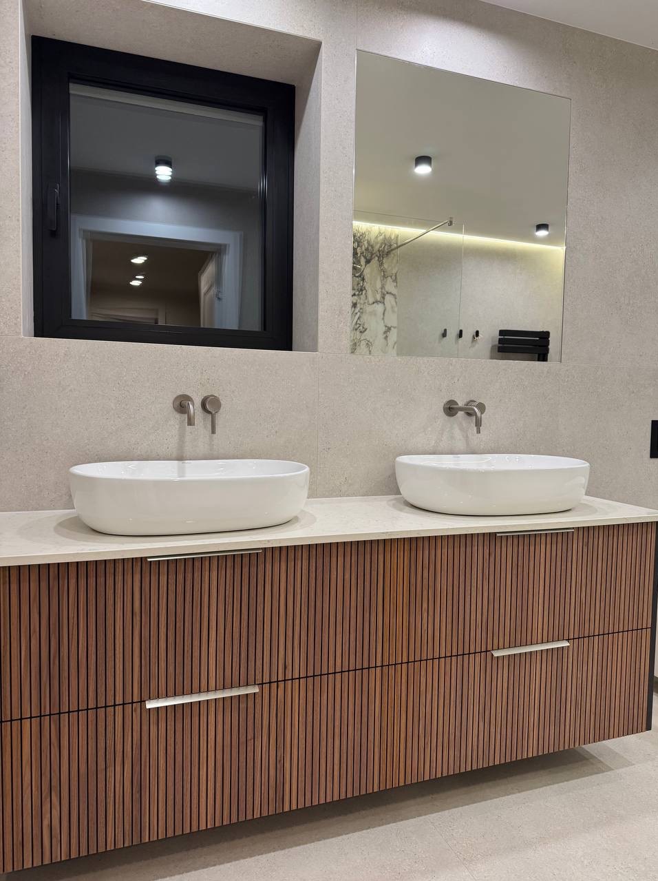 Nowoczesna łazienka z dwoma białymi umywalkami nablatowymi, armaturą w kolorze stali szczotkowanej, szafką z drewnianymi lamelami i czarną ramą okna.