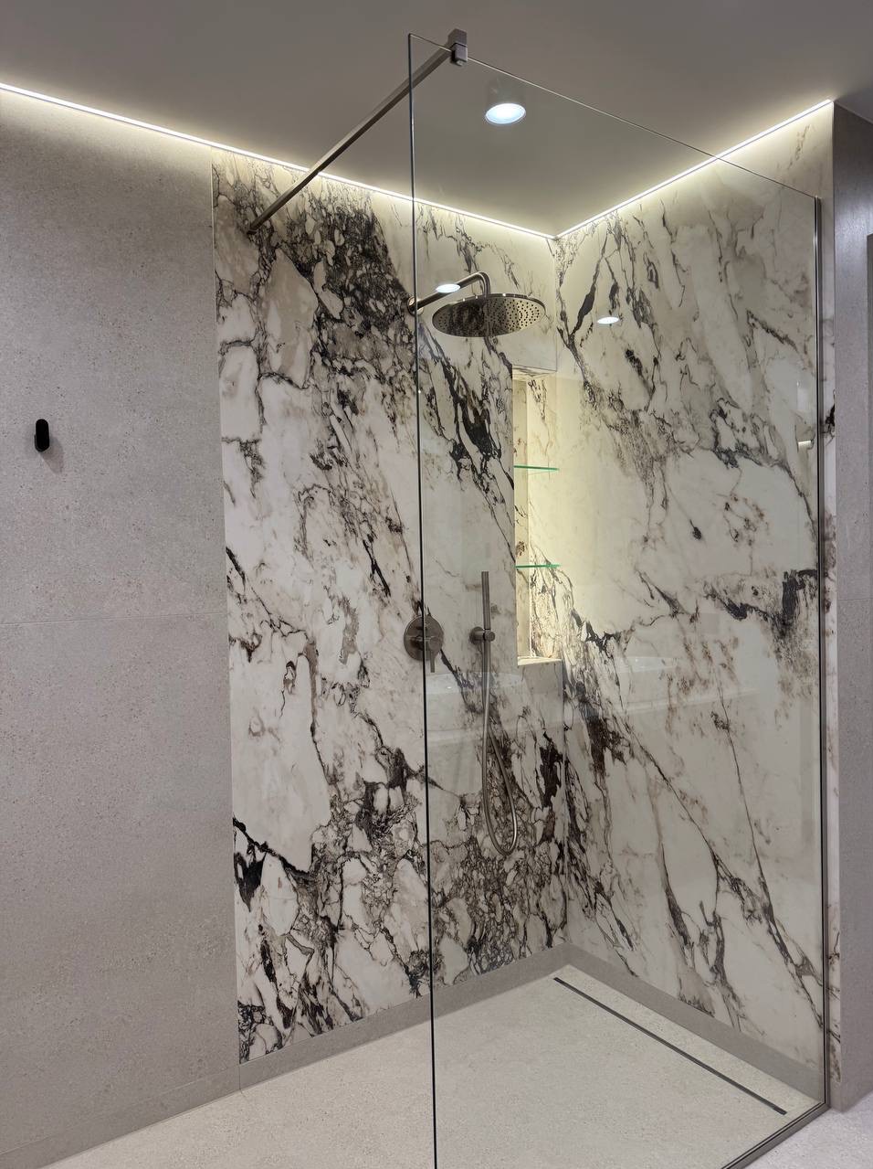Nowoczesna kabina prysznicowa z marmurowymi płytami, szklaną ścianką i podświetleniem LED, widoczna półka wnękowa.