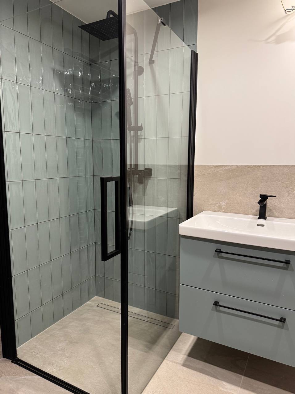 Nowoczesna łazienka po remoncie: kabina prysznicowa typu walk-in z czarną ramą i deszczownicą, umywalka z szafką w pastelowym odcieniu niebieskiego, czarna bateria.