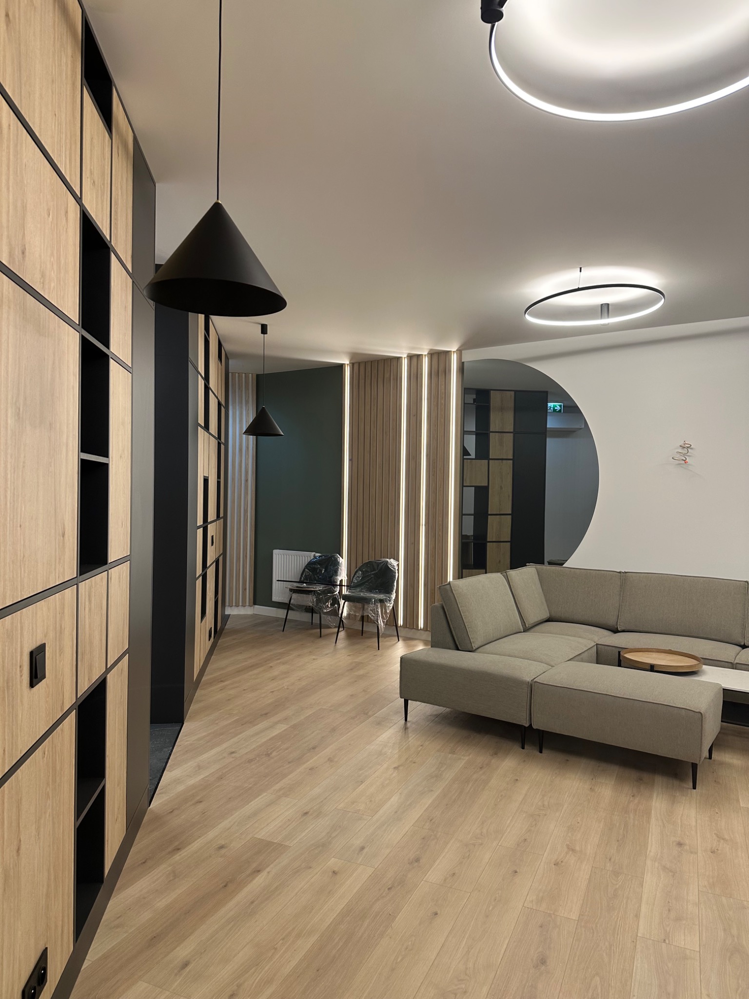 Nowoczesne wnętrze biurowe z drewnianą zabudową, szarą sofą narożną i okrągłymi lampami wiszącymi, akcentowane czarnymi elementami wykończeniowymi i podłogą w jasnym drewnie.
