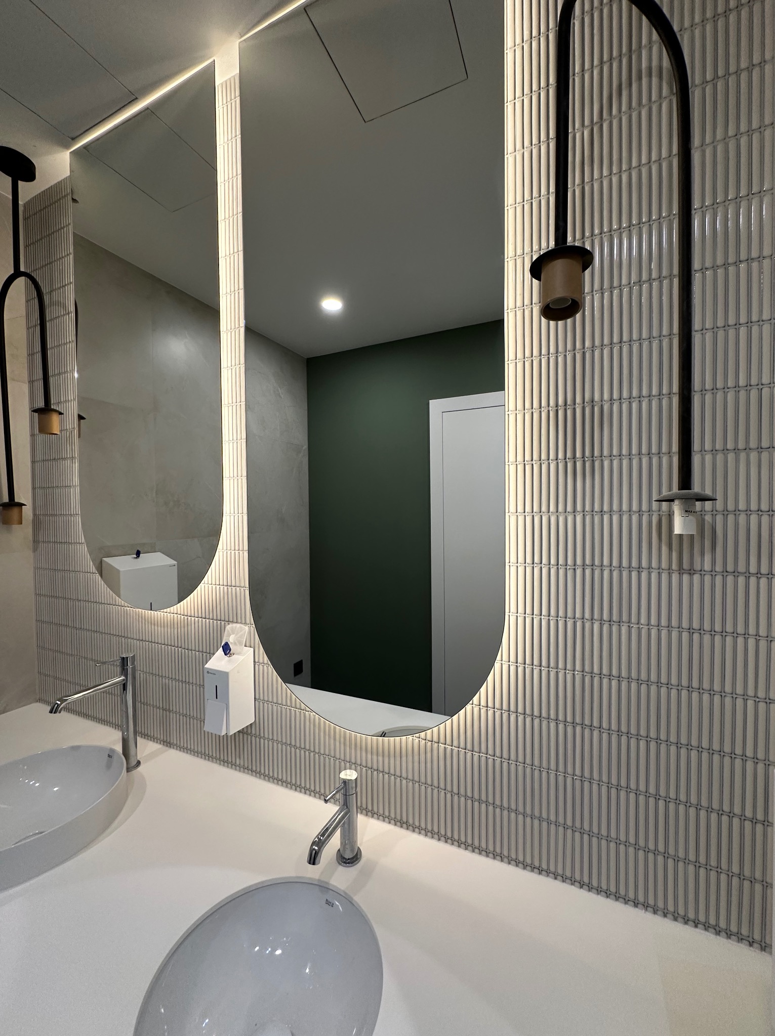 Nowoczesna łazienka z dwoma owalnymi umywalkami nablatowymi, podświetlanym lustrem w kształcie łuku i szarą, pionową mozaiką na ścianie, uzupełniona czarnymi lampami w stylu industrialnym.