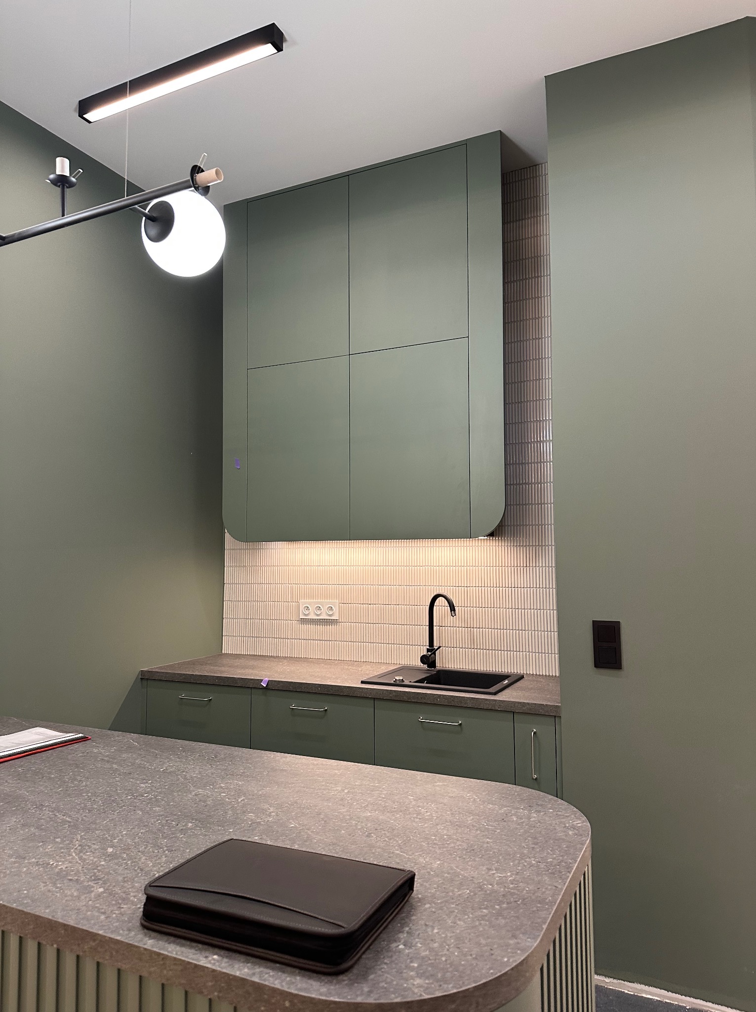 Nowoczesna kuchnia w odcieniach zieleni z minimalistyczną zabudową, czarną armaturą i blatem z szarego kamienia, oświetlona lampą sufitową z kulistym kloszem.