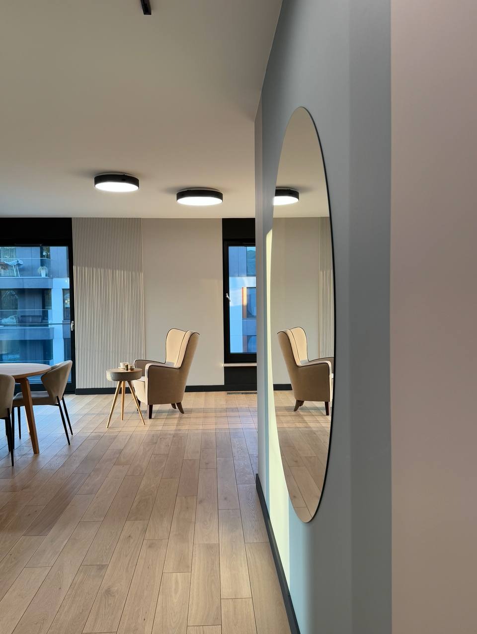 Jasne wnętrze salonu z parkietem, beżowymi fotelami i okrągłym lustrem na ścianie, odbijającym przestrzeń pokoju.