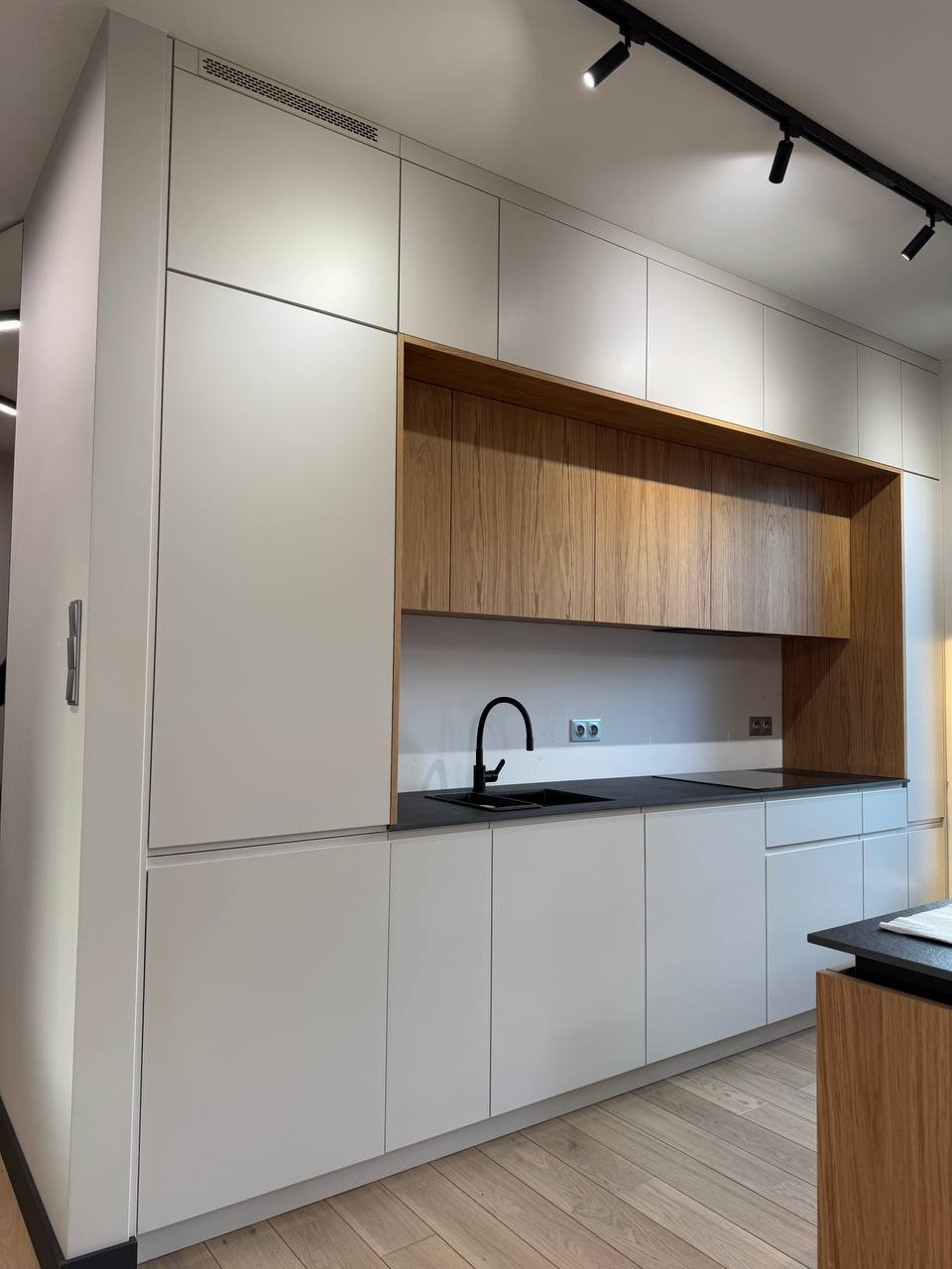 Nowoczesna kuchnia z białymi szafkami bez uchwytów, drewnianą zabudową nad blatem, czarnym zlewem i baterią oraz oświetleniem szynowym na suficie.