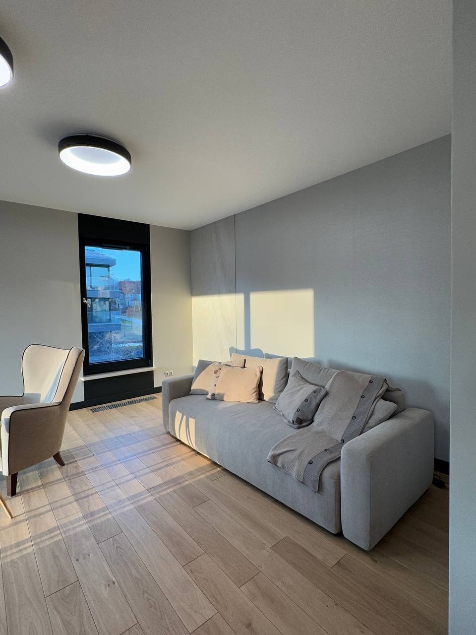 Jasne, przestronne wnętrze salonu z drewnianą podłogą, szarą sofą z poduszkami i pledem, oraz kremowym fotelem uszakiem, oświetlone popołudniowym słońcem wpadającym przez okno z czarną ramą.