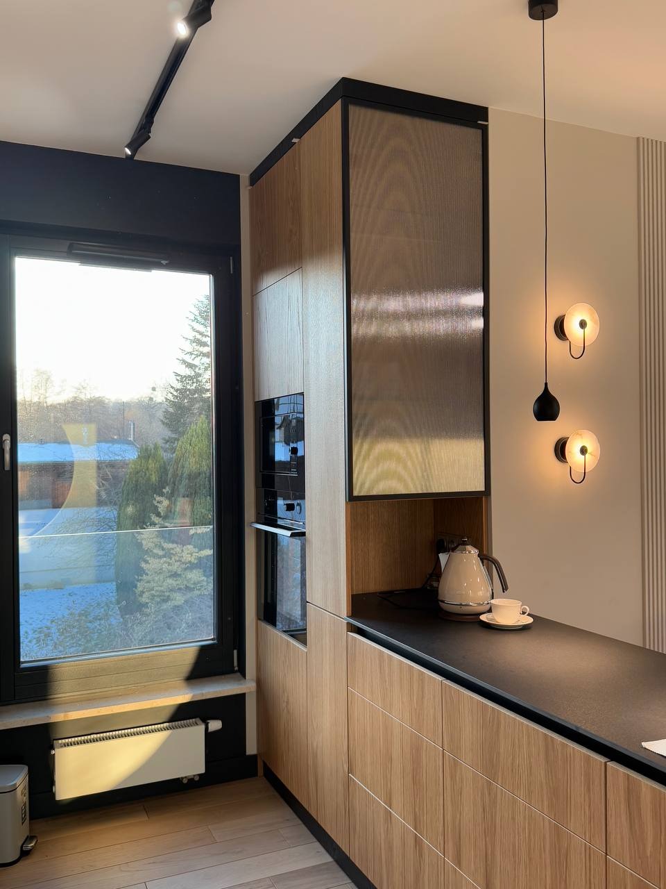 Nowoczesna kuchnia z drewnianymi frontami, czarnym blatem i oknem z widokiem na zimowy krajobraz, uzupełniona o designerskie lampy ścienne i czajnik z filiżanką.