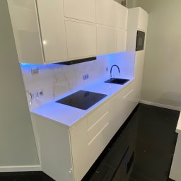 Wood House Tomasz Gaj - Biała, nowoczesna kuchnia z podświetleniem LED, czarnym blatem i płytą indukcyjną. Minimalistyczny design, błyszczące fronty szafek, czarne płytki podłogowe.