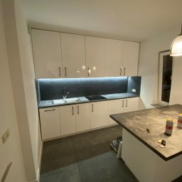 Wood House Tomasz Gaj - Nowoczesna, biała kuchnia na wymiar z czarnym blatem i oświetleniem LED w Witoldzinie. Minimalistyczny design, widoczne puszki na blacie dodają realizmu.