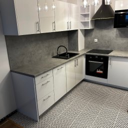Wood House Tomasz Gaj - Biała, nowoczesna kuchnia z szarym blatem i wzorzystą podłogą. Widoczne szafki wiszące z oświetleniem, piekarnik i płyta indukcyjna. Minimalistyczny design.