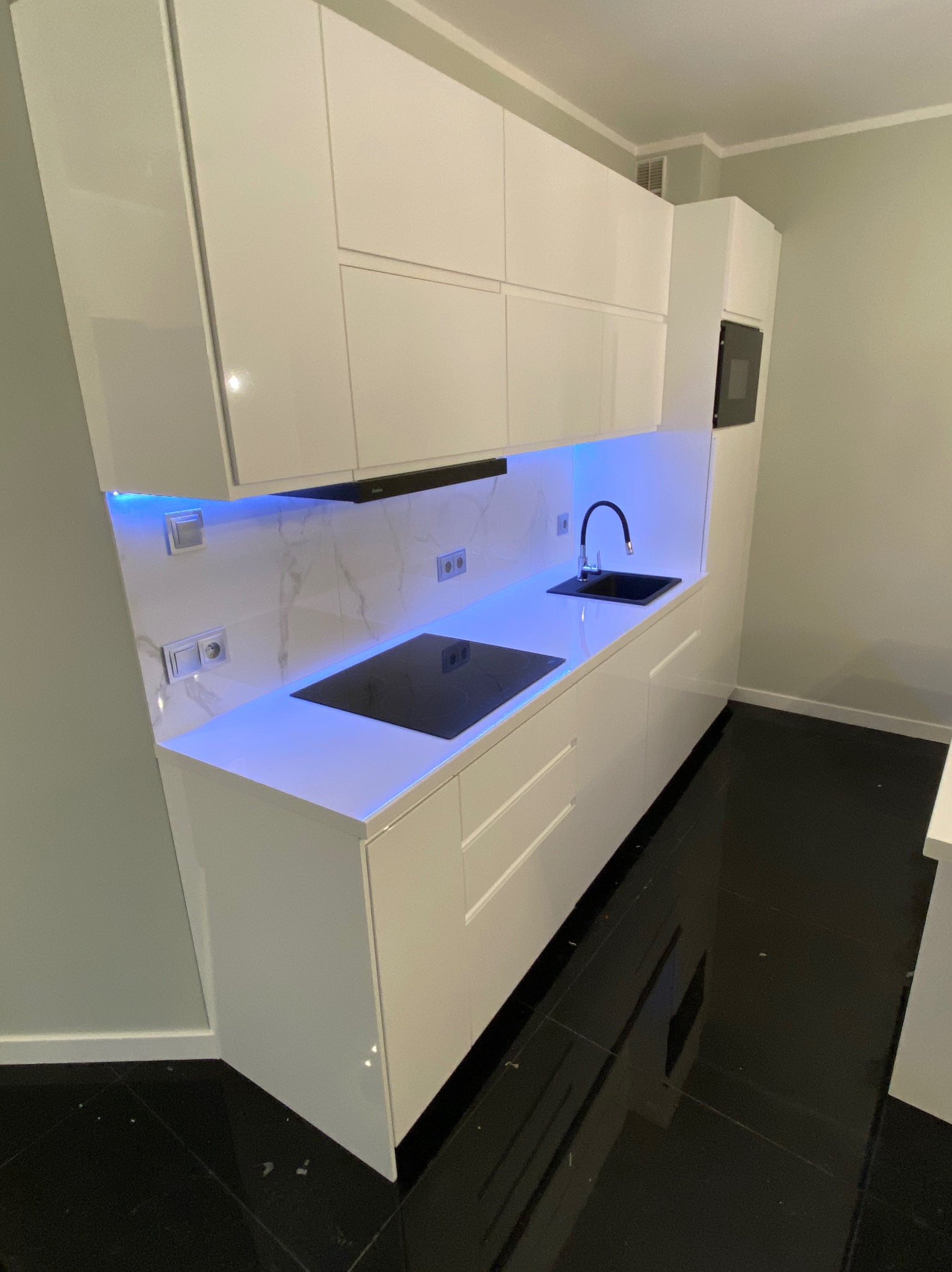 Biała, nowoczesna kuchnia z podświetleniem LED, czarnym blatem i płytą indukcyjną. Minimalistyczny design, błyszczące fronty szafek, czarne płytki podłogowe.