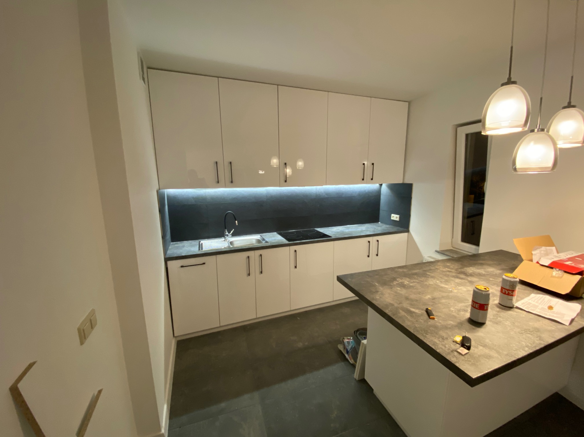 Nowoczesna, biała kuchnia na wymiar z czarnym blatem i oświetleniem LED w Witoldzinie. Minimalistyczny design, widoczne puszki na blacie dodają realizmu.