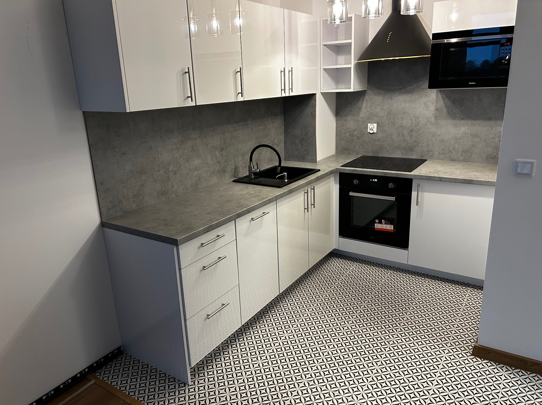 Biała, nowoczesna kuchnia z szarym blatem i wzorzystą podłogą. Widoczne szafki wiszące z oświetleniem, piekarnik i płyta indukcyjna. Minimalistyczny design.