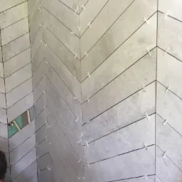 Ściana w trakcie układania płytek w jodełkę, z użyciem krzyżyków dystansowych, widoczny fragment ściany bez płytek z zieloną płytą gipsową.