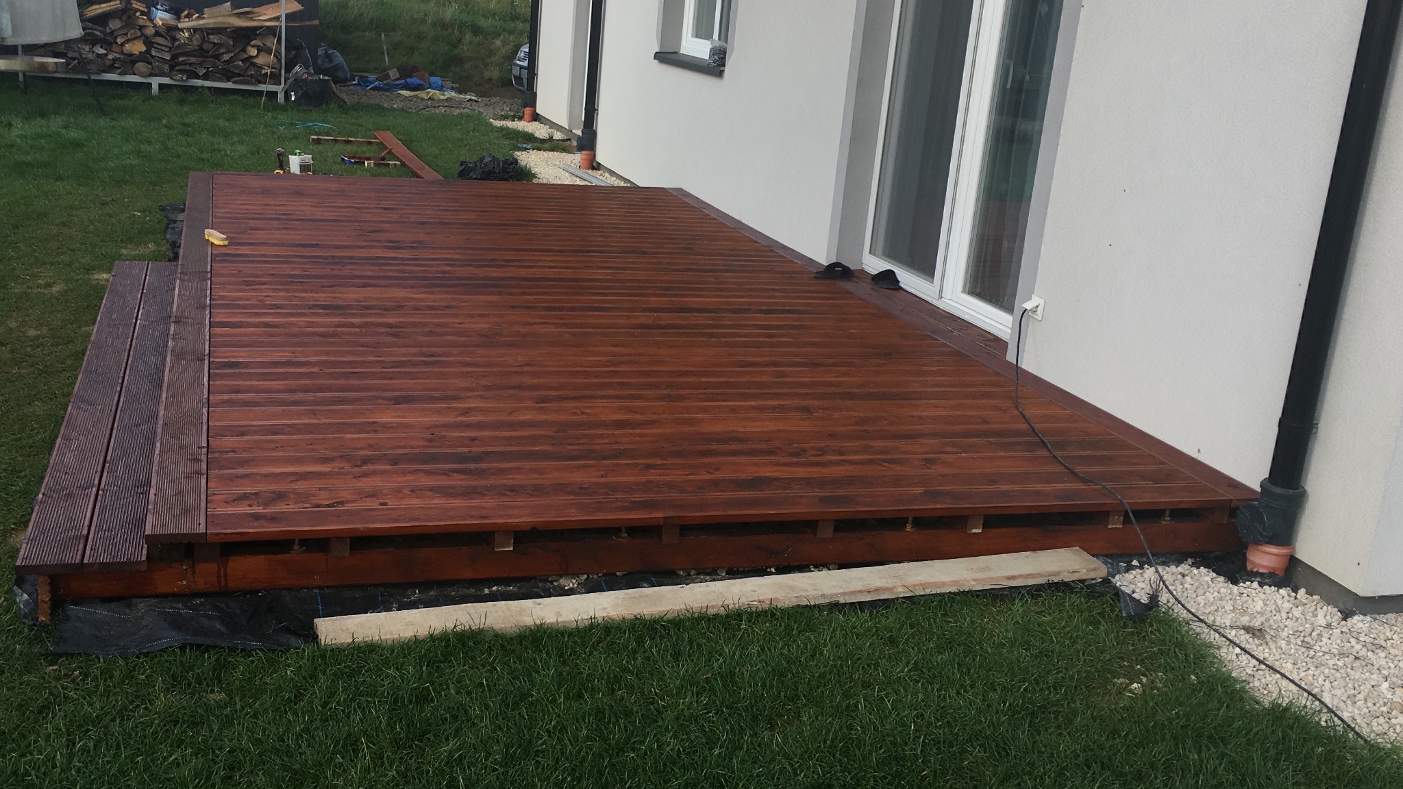 Świeżo zaimpregnowany taras z drewna egzotycznego z dwustopniowymi schodami, przylegający do białego budynku z oknami, otoczony zielonym trawnikiem i częściowo widocznym stosem drewna.