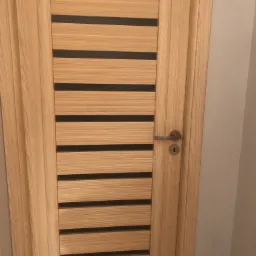 Jasne, drewniane drzwi wewnętrzne z poziomymi, czarnymi paskami i srebrną klamką na tle szarej ściany.