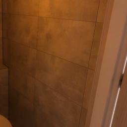 Maciej i spółka - Fragment ściany łazienki wyłożonej brązowymi, matowymi płytkami ceramicznymi, widoczny fragment toalety i ościeżnica drzwi